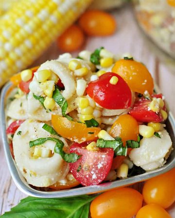 Tortellini Pasta Salad in a bowl with corn on the cob, colorful grape tomatoes, and basil leaf in background